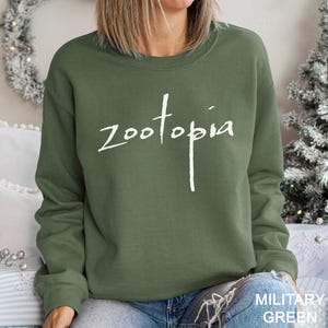 May include: Olive green sweatshirt with the word "Zootopia" in white script. The sweatshirt is a solid color and has a crew neck. The text "MILITARY GREEN" is in the bottom right corner.