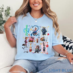 May include: Light blue t-shirt featuring a colorful graphic of characters from the animated film Zootopia. The design includes various animal characters in a grid layout, with the film's title in green. The shirt also has the text "LIGHT BLUE" at the bottom.