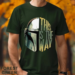 Pu&ograve; includere: Una t-shirt a maniche corte color verde bosco con una stampa grafica di un casco metallico sul lato sinistro e il testo THIS IS THE WAY in lettere maiuscole gialle e verdi sul lato destro, centrato sul petto del capo di abbigliamento.
