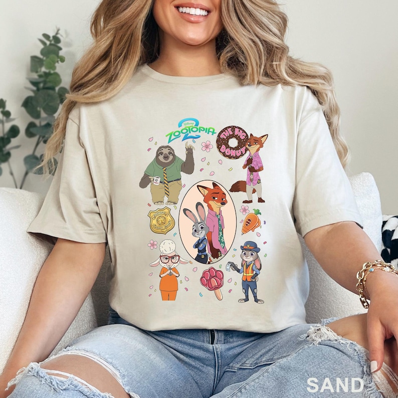 K&ouml;nnte beinhalten: Ein sandfarbenes T-Shirt mit einem Zootopia 2 Grafikdruck. Das Design zeigt Charaktere wie Nick Wilde, Judy Hopps, Flash und Bellwether sowie Symbole wie einen Donut, eine Karotte, ein Abzeichen und ein pfotenf&ouml;rmiges Eis am Stiel.