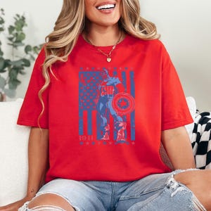 May include: Red t-shirt featuring a distressed graphic of Captain America holding his shield in front of an American flag design. The graphic is in blue and white, with the year "1941" at the bottom. The shirt is a casual, oversized fit.