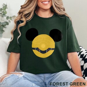 May include: Forest green t-shirt featuring a gold and black graphic design. The design includes a yellow circle with black ears and a detailed face. The text "FOREST GREEN" is printed at the bottom of the shirt.