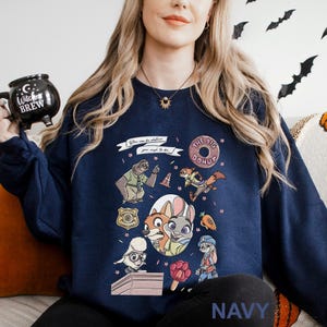 May include: Navy blue sweatshirt featuring a colorful graphic of characters from a popular animated movie. The design includes a sloth, fox, bunny, and other animals, along with the text "You can be whatever you wish to be." The word "NAVY" is printed at the bottom.