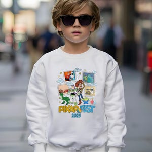 Może przedstawiać: Biała bluza z kolorową grafiką postaci z filmu Pixar, w tym Woody'ego i Buzza Astrala. Projekt zawiera napis "Pixar Fest 2025". Bluza jest noszona przez osobę w okularach przeciwsłonecznych.