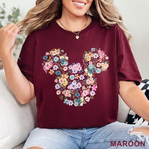 May include: Maroon t-shirt featuring a floral Mickey Mouse design. The design is made up of colorful flowers in shades of pink, blue, yellow, and white. The word "MAROON" is printed at the bottom right corner of the shirt.