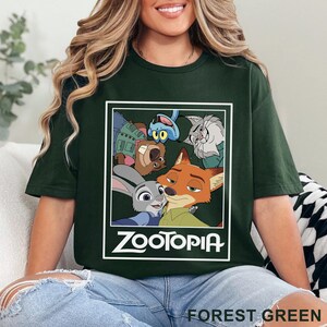 May include: Forest green t-shirt with a graphic of characters from the animated film Zootopia. The design includes Judy Hopps, Nick Wilde, and other characters within a white rectangular frame. The word "ZOOTOPIA" is printed in white.