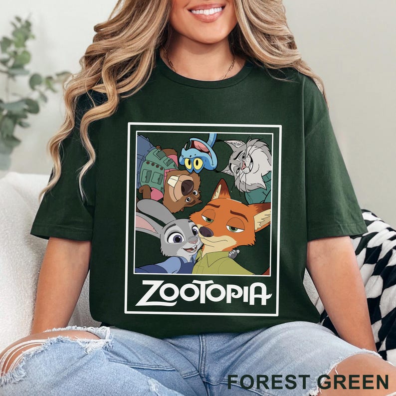 May include: Forest green t-shirt with a graphic of characters from the animated film Zootopia. The design includes Judy Hopps, Nick Wilde, and other characters within a white rectangular frame. The word "ZOOTOPIA" is printed in white.