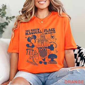 May include: Orange t-shirt with a navy blue graphic design featuring Disney-themed icons and text. The text reads "The Most Magical Place on Earth" and includes images of Mickey Mouse ears, a castle, and other park-related elements.
