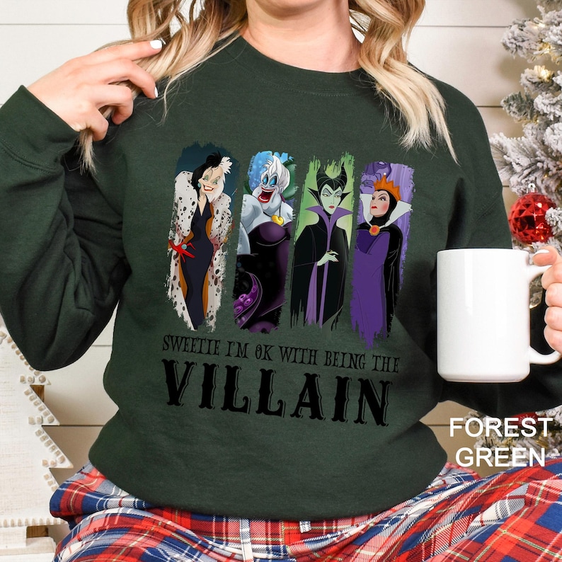 Puede incluir: Sudadera verde bosque con un dise&ntilde;o de cuatro villanos de dibujos animados. El texto "Sweetie I'm ok with being the Villain" est&aacute; impreso debajo de los personajes. Una taza blanca se sostiene en la mano.