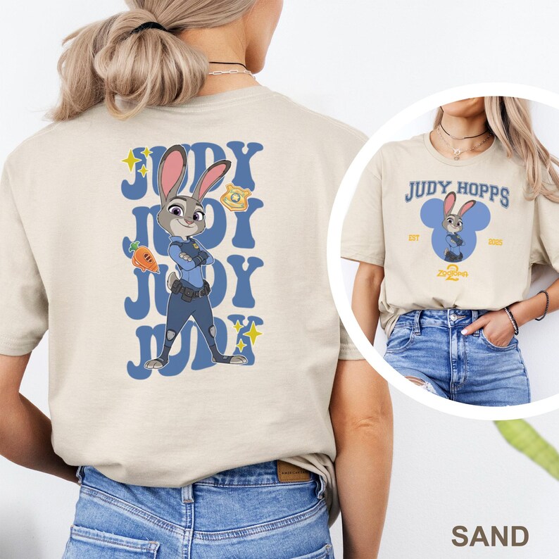 May include: Sand-colored t-shirt featuring a cartoon image of Judy Hopps from Zootopia. The back of the shirt has a blue design with the name "JUDY" repeated, and an image of Judy Hopps holding a carrot. The front of the shirt has the text "JUDY HOPPS" and a cartoon image of Judy.