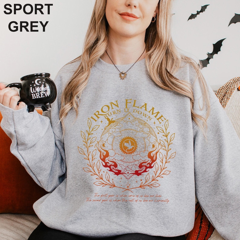 May include: Sport grey sweatshirt with a circular graphic that reads "IRON FLAME BURN IT DOWN" in gold and red. The sweatshirt also has text at the bottom. A black mug with "Witches Brew" is held in the image.
