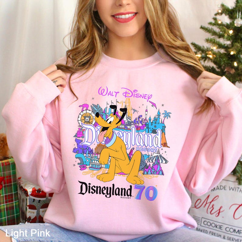 May include: Light pink sweatshirt featuring a colorful Disneyland graphic with Pluto and the text "Disneyland 70". The design includes iconic park elements and the words "Walt Disney Resort".