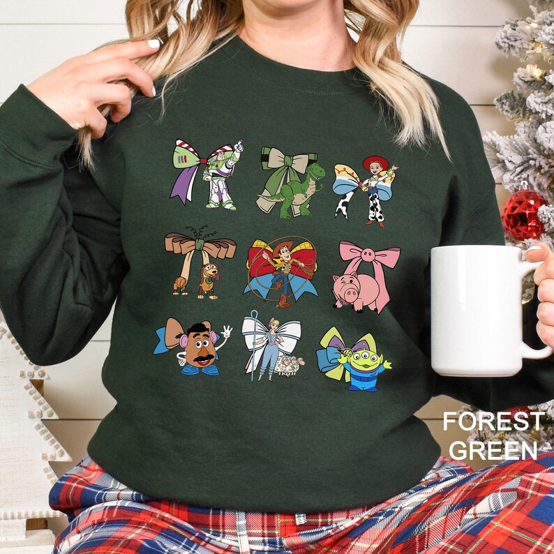 May include: Forest green sweatshirt featuring cartoon character bows. The bows are designed with characters like Buzz Lightyear, Rex, Jessie, and others. The text "FOREST GREEN" is printed on the bottom right.