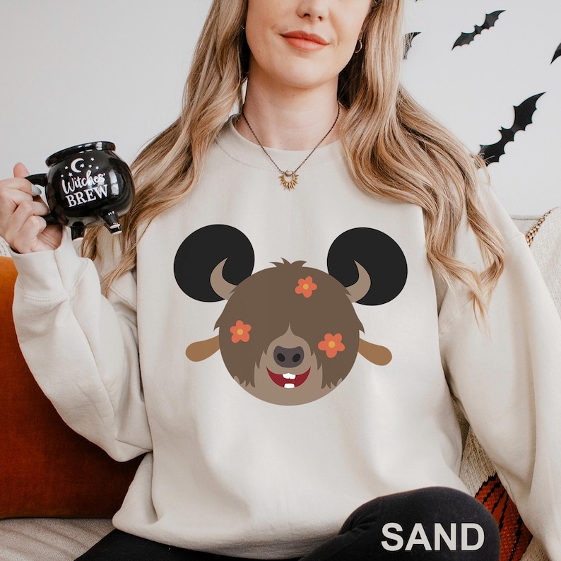 May include: Sand-colored sweatshirt featuring a cartoon bear face with large black ears, brown fur, and orange flower accents. The bear has a smiling expression with two white teeth. The word "SAND" is printed at the bottom.