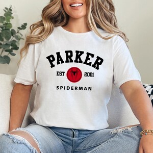 May include: White t-shirt with the text "PARKER EST 2001 SPIDERMAN" in black and red. The design includes a red circle with a spider symbol. The shirt is a casual wear item.