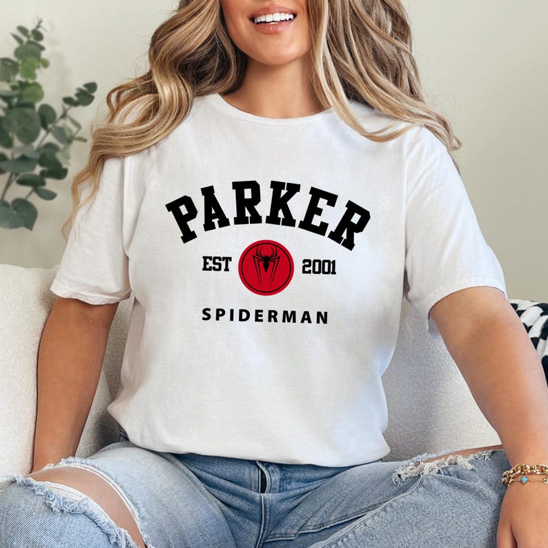 May include: White t-shirt with the text "PARKER EST 2001 SPIDERMAN" in black and red. The design includes a red circle with a spider symbol. The shirt is a casual wear item.