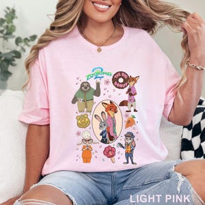 K&ouml;nnte beinhalten: Ein hellrosa T-Shirt mit einem Zootopia-Grafikdruck, der Charaktere wie Judy Hopps, Nick Wilde, Flash das Faultier und Bellwether zeigt. Das Design enth&auml;lt das Zootopia 2-Logo, einen Donut, eine Karotte, ein Pfoten-Eis am Stiel und ein Polizeimarken-Symbol.