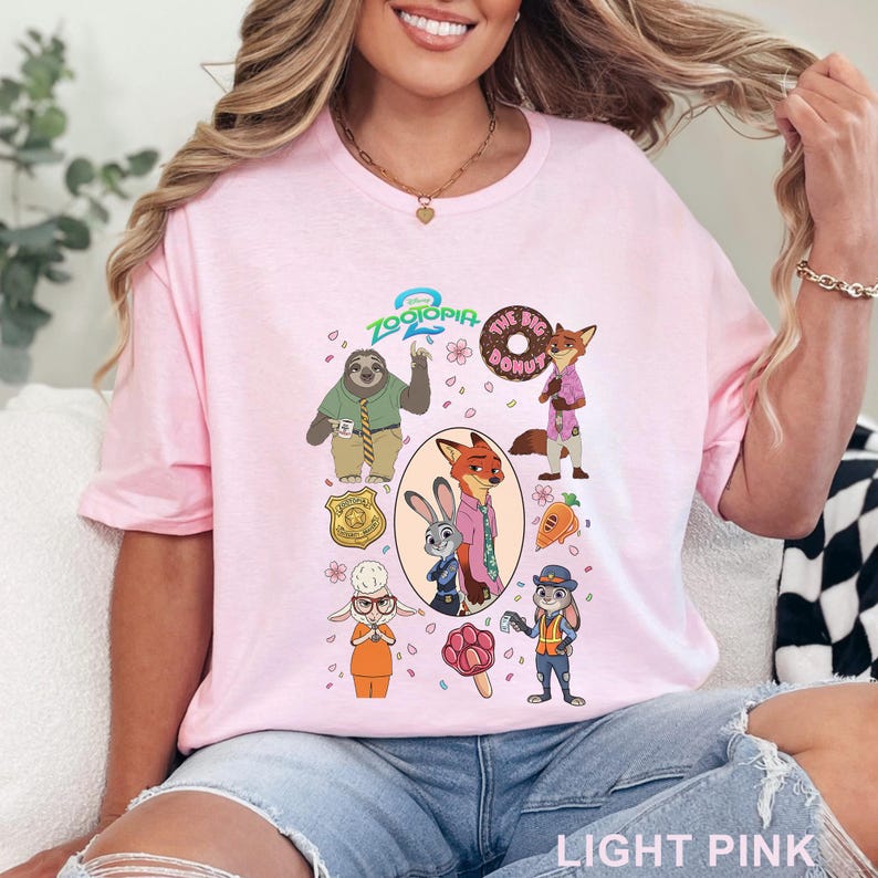 K&ouml;nnte beinhalten: Ein hellrosa T-Shirt mit einem Zootopia-Grafikdruck, der Charaktere wie Judy Hopps, Nick Wilde, Flash das Faultier und Bellwether zeigt. Das Design enth&auml;lt das Zootopia 2-Logo, einen Donut, eine Karotte, ein Pfoten-Eis am Stiel und ein Polizeimarken-Symbol.