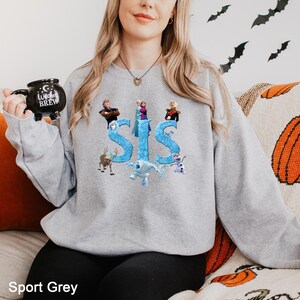 K&ouml;nnte beinhalten: Ein hellgraues Sweatshirt mit Rundhalsausschnitt und einem Grafikdruck des Wortes SIS in einer blauen, eisigen Schriftart, umgeben von Charakteren aus dem Film Frozen, darunter Elsa, Anna, Kristoff, Olaf, Sven und Marshmallow, mit einer schwarzen Tasse.