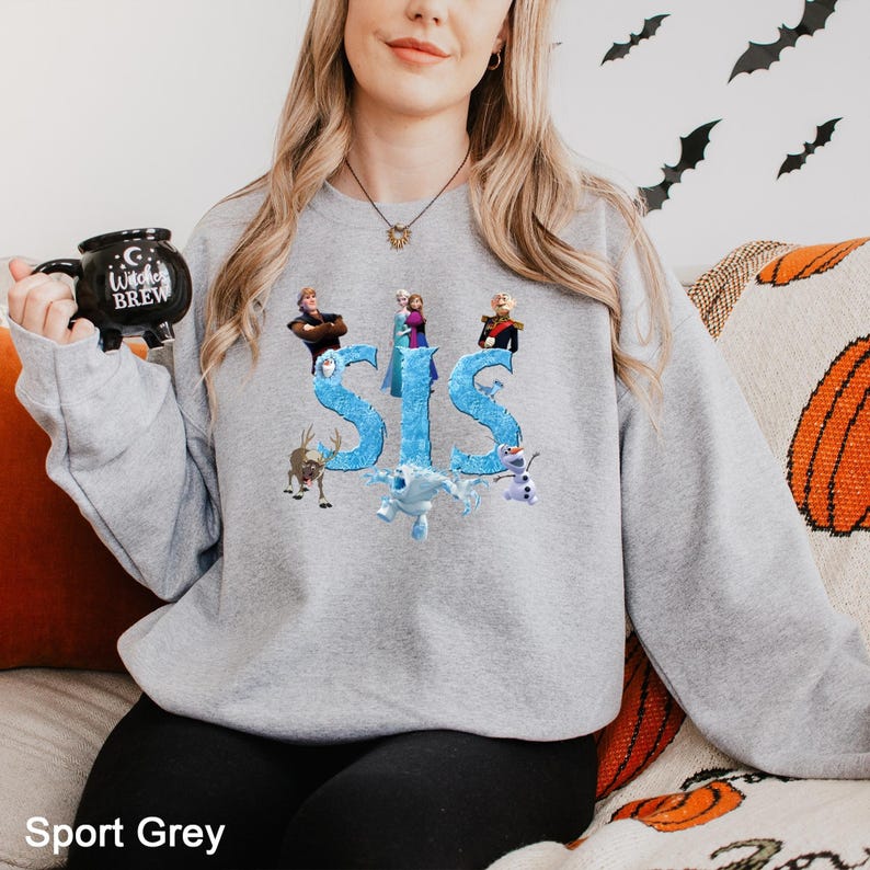 K&ouml;nnte beinhalten: Ein hellgraues Sweatshirt mit Rundhalsausschnitt und einem Grafikdruck des Wortes SIS in einer blauen, eisigen Schriftart, umgeben von Charakteren aus dem Film Frozen, darunter Elsa, Anna, Kristoff, Olaf, Sven und Marshmallow, mit einer schwarzen Tasse.