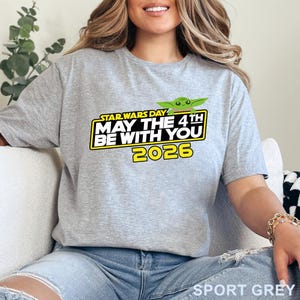 May include: A light gray t-shirt with the text "STAR WARS DAY MAY THE 4TH BE WITH YOU 2026" in yellow and white, featuring a green cartoon character. The shirt is made of a soft material.
