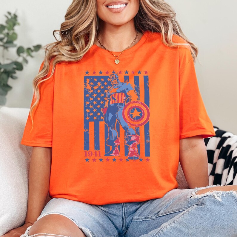 May include: Orange t-shirt featuring a Captain America graphic with a shield and the American flag. The design includes the year 1941 and stars. The shirt is a vibrant orange color.