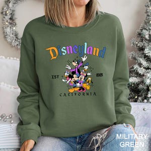 Puede incluir: Sudadera verde militar con el logotipo de Disneyland en naranja y azul, con un gr&aacute;fico de Mickey Mouse, Minnie Mouse, Donald Duck, Goofy y Pluto. El texto "EST 2025 CALIFORNIA" tambi&eacute;n est&aacute; impreso en la sudadera.