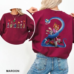 May include: A maroon crewneck sweatshirt featuring a large graphic on the back of a blue snake coiled around a fox and a rabbit. A circular inset shows the front of the sweatshirt with a smaller graphic of multiple characters holding balloons.