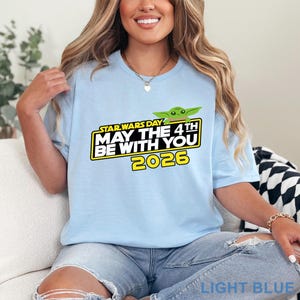 May include: Light blue t-shirt featuring a Star Wars Day graphic with the text "May the 4th Be With You 2026" and a green Baby Yoda illustration. The shirt is a light blue color.