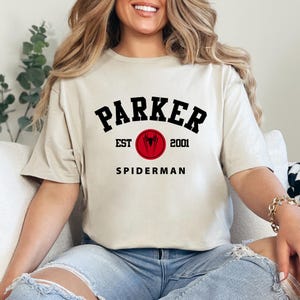 May include: Beige t-shirt with the text "PARKER" in black, arched above "SPIDERMAN". A red circle with a spider symbol and the text "EST 2001" is centered below "PARKER". The shirt is a casual, short-sleeved style.