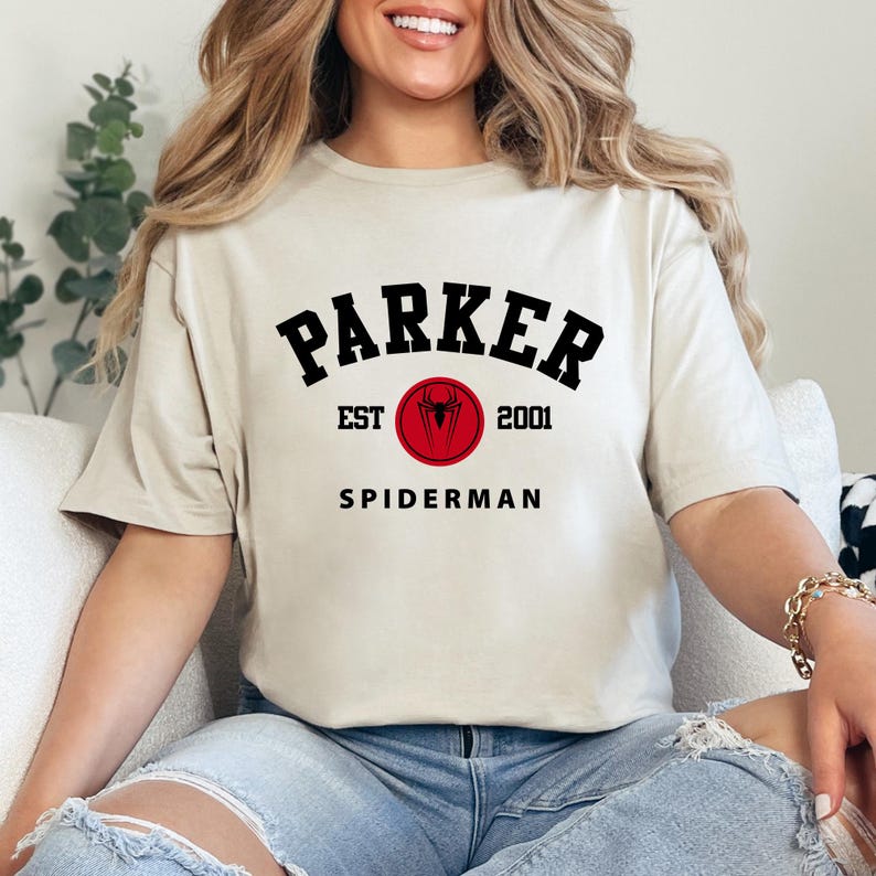 May include: Beige t-shirt with the text "PARKER" in black, arched above "SPIDERMAN". A red circle with a spider symbol and the text "EST 2001" is centered below "PARKER". The shirt is a casual, short-sleeved style.