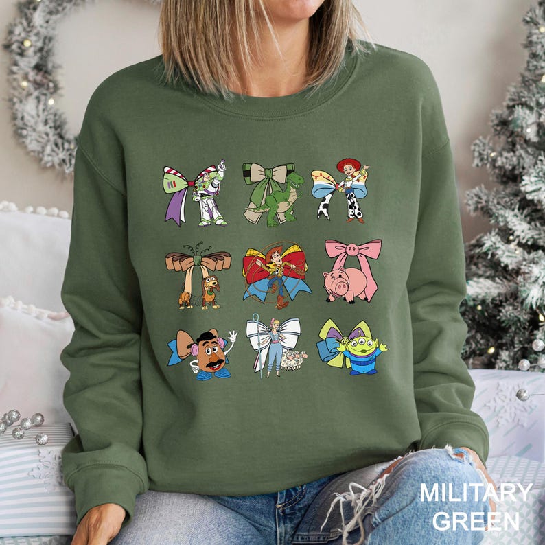 May include: Military green sweatshirt featuring colorful cartoon character bow designs. The bows include characters like Buzz Lightyear, Woody, and others. The text "MILITARY GREEN" is printed at the bottom right.