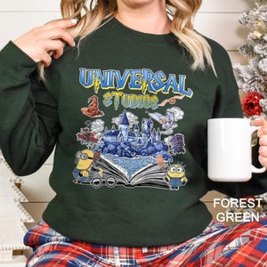 Op de afbeelding: Bostgroen sweatshirt met een Universal Studios-afbeelding met een kasteel, Minions en tekst. Het ontwerp bevat een tovenaars hoed, een uil en een open boek. Het sweatshirt wordt gecombineerd met een geruite broek.
