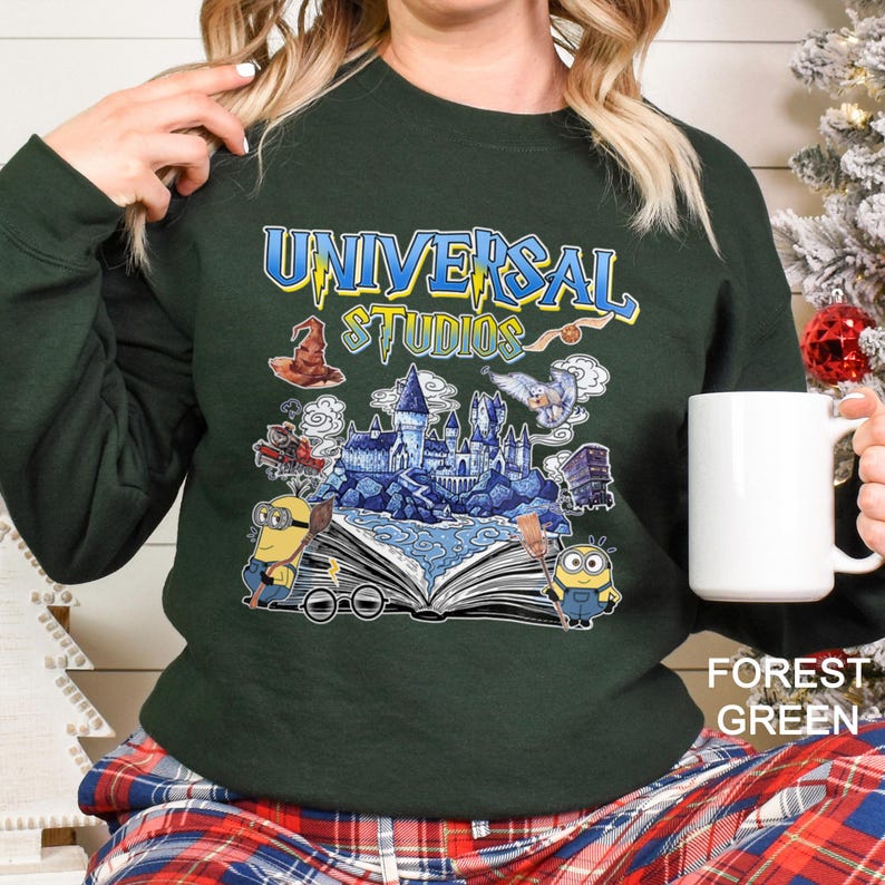 Op de afbeelding: Bostgroen sweatshirt met een Universal Studios-afbeelding met een kasteel, Minions en tekst. Het ontwerp bevat een tovenaars hoed, een uil en een open boek. Het sweatshirt wordt gecombineerd met een geruite broek.
