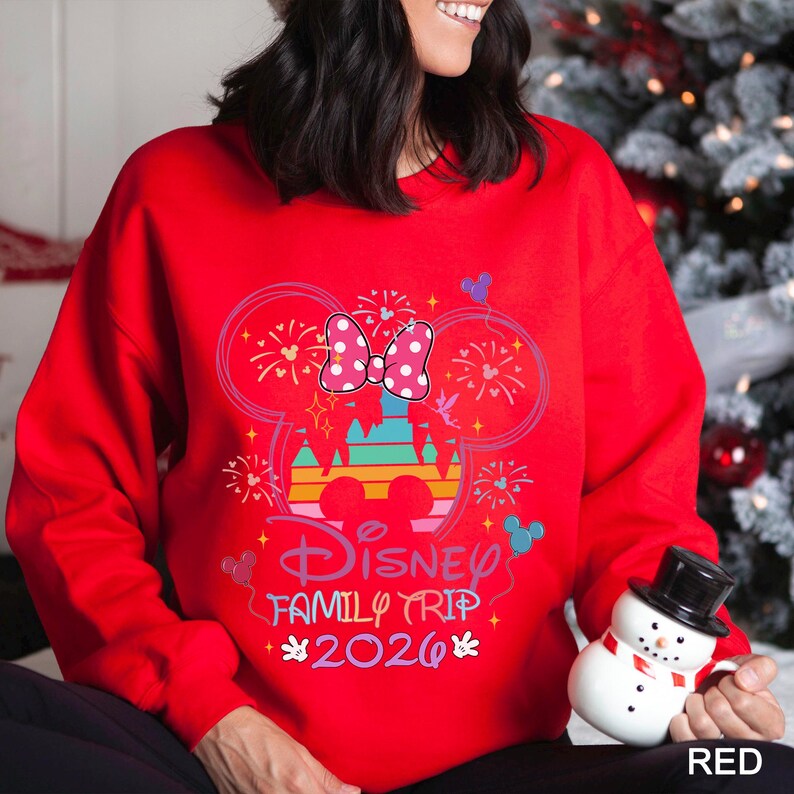 May include: A red sweatshirt features a colorful graphic of the Disney logo with fireworks, a bow, and the text "Disney Family Trip 2026". The design includes a castle silhouette and Mickey Mouse ears.