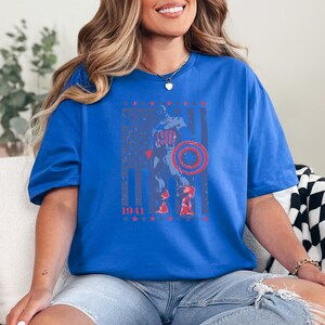 May include: A blue t-shirt featuring a graphic of Captain America with a shield, set against a distressed American flag design. The design includes the year "1941" and red and blue stars.