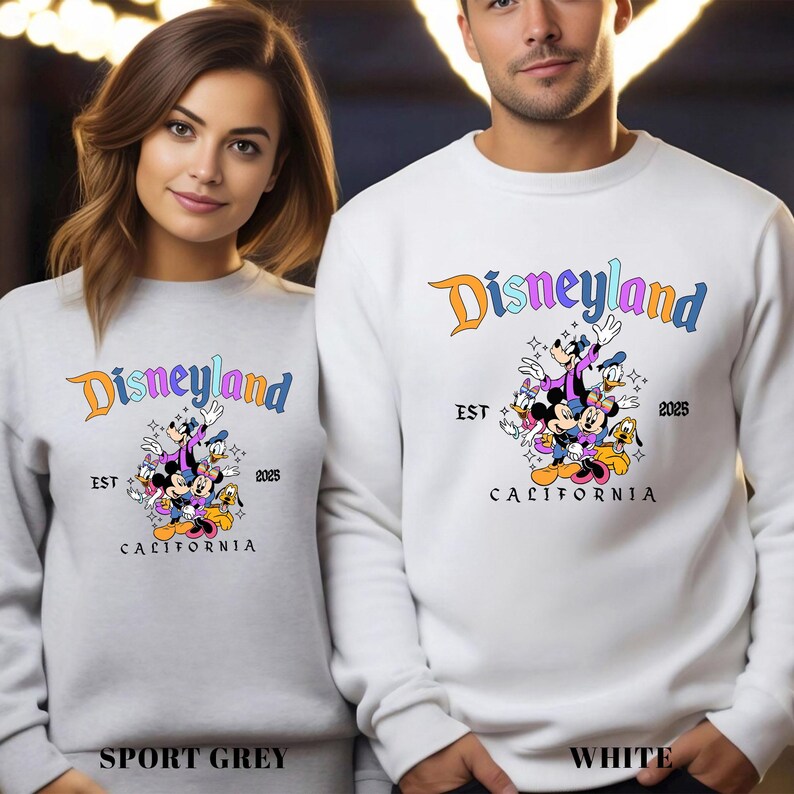 Puede incluir: Sudaderas grises deportivas y blancas con el logotipo de Disneyland y varios personajes de dibujos animados. Las sudaderas tienen el texto "EST 2025 CALIFORNIA". El logotipo de Disneyland es naranja, morado y azul.