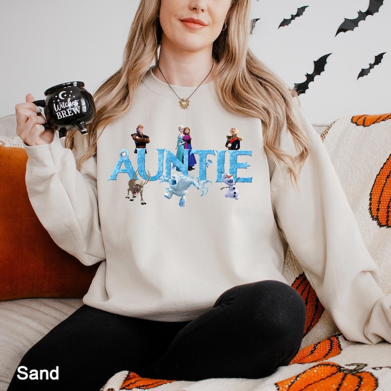 K&ouml;nnte beinhalten: Ein sandfarbenes Sweatshirt mit Rundhalsausschnitt, das das Wort AUNTIE in einer blauen, eisigen Schriftart zeigt, verziert mit verschiedenen animierten Charakteren aus der Frozen-Filmreihe. Das Sweatshirt wird mit einem schwarzen Kesselbecher kombiniert.