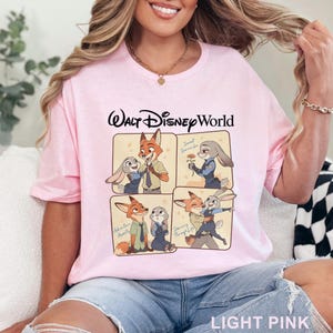 May include: Light pink t-shirt with "Walt Disney World" text and cartoon images of Nick Wilde and Judy Hopps from Zootopia. The shirt features four panels with different scenes of the characters. The words "Sweet Surrender", "Adventures Await", and "Dancing Through Life" are also visible.