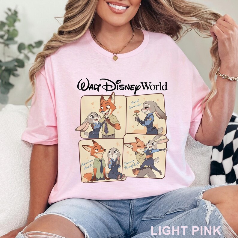 May include: Light pink t-shirt with "Walt Disney World" text and cartoon images of Nick Wilde and Judy Hopps from Zootopia. The shirt features four panels with different scenes of the characters. The words "Sweet Surrender", "Adventures Await", and "Dancing Through Life" are also visible.