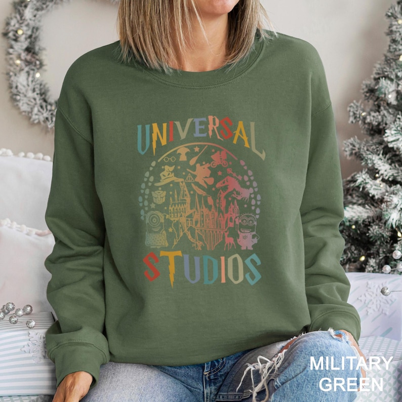 May include: Olive green sweatshirt with the words "UNIVERSAL STUDIOS" in a colorful, vintage-style design. The graphic includes various characters and imagery associated with Universal Studios.