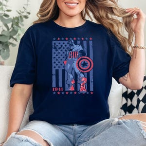 May include: Navy blue t-shirt featuring a Captain America graphic. The design includes the American flag, the year "1941", and a depiction of Captain America with his shield. The graphic is in shades of red, white, and blue.