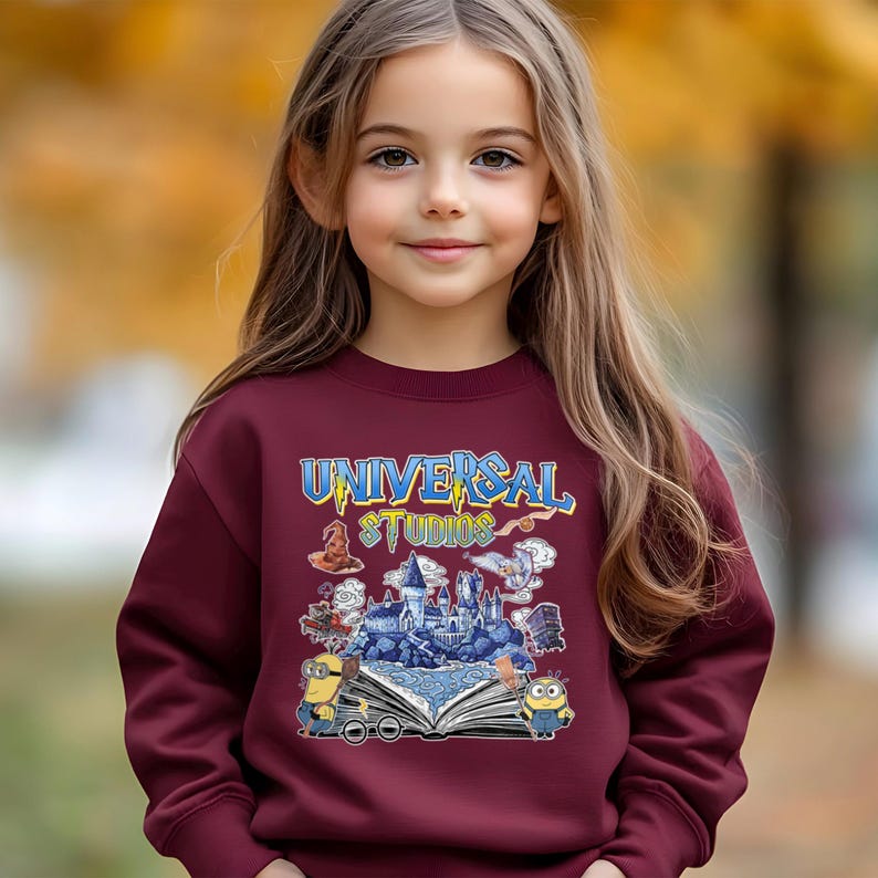 Op de afbeelding: Een bordeauxrode sweatshirt met de tekst "UNIVERSAL STUDIOS" in blauw en geel. De afbeelding bevat illustraties van personages en sc&egrave;nes uit Universal Studios-films, waaronder Minions en een kasteel.