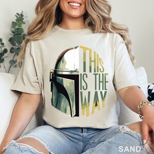Pu&ograve; includere: Una t-shirt color sabbia con una stampa grafica di un casco mandaloriano sul lato sinistro e il testo THIS IS THE WAY in lettere maiuscole verdi e gialle sul lato destro, il tutto su un motivo di sfondo circolare.
