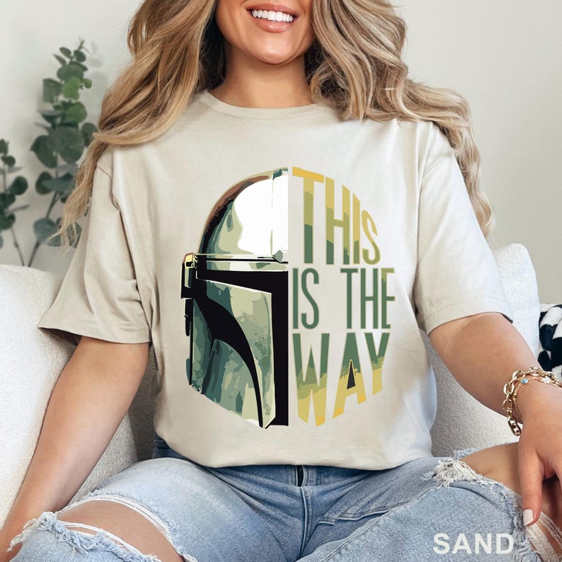 Pu&ograve; includere: Una t-shirt color sabbia con una stampa grafica di un casco mandaloriano sul lato sinistro e il testo THIS IS THE WAY in lettere maiuscole verdi e gialle sul lato destro, il tutto su un motivo di sfondo circolare.