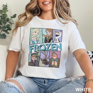 May include: White t-shirt featuring a colorful graphic of characters from the animated film "Frozen." The design includes Elsa, Anna, Olaf, Kristoff, and Sven, with the word "FROZEN" in a blue and teal font. The shirt is made of a soft material.
