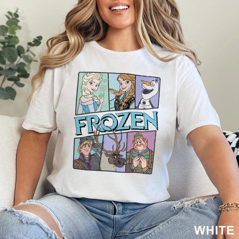 May include: White t-shirt featuring a colorful graphic of characters from the animated film "Frozen." The design includes Elsa, Anna, Olaf, Kristoff, and Sven, with the word "FROZEN" in a blue and teal font. The shirt is made of a soft material.