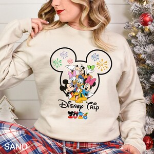 May include: Sand-colored sweatshirt featuring a black Mickey Mouse head outline with colorful fireworks and Disney characters. The text "Disney Trip 2023" is printed below the characters. The sweatshirt is long-sleeved.