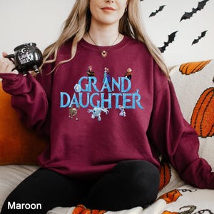 K&ouml;nnte beinhalten: Ein kastanienbraunes Langarm-Sweatshirt mit dem Text GRAND DAUGHTER in blauen Buchstaben, umgeben von verschiedenen animierten Figuren. Das Sweatshirt wird mit einer schwarzen Hose und einer schwarzen Tasse mit dem Aufdruck Witches Brew getragen.