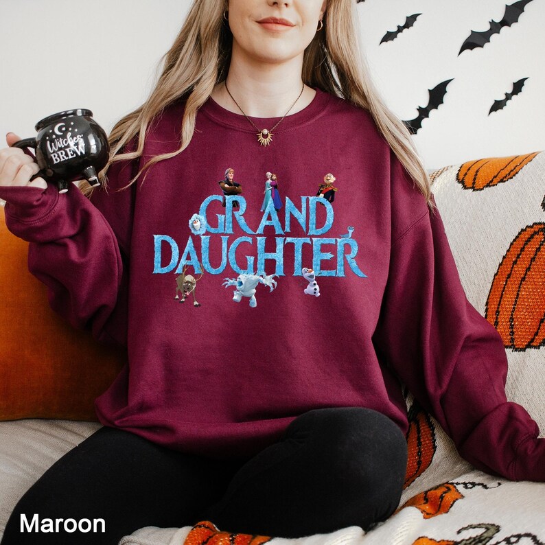 K&ouml;nnte beinhalten: Ein kastanienbraunes Langarm-Sweatshirt mit dem Text GRAND DAUGHTER in blauen Buchstaben, umgeben von verschiedenen animierten Figuren. Das Sweatshirt wird mit einer schwarzen Hose und einer schwarzen Tasse mit dem Aufdruck Witches Brew getragen.